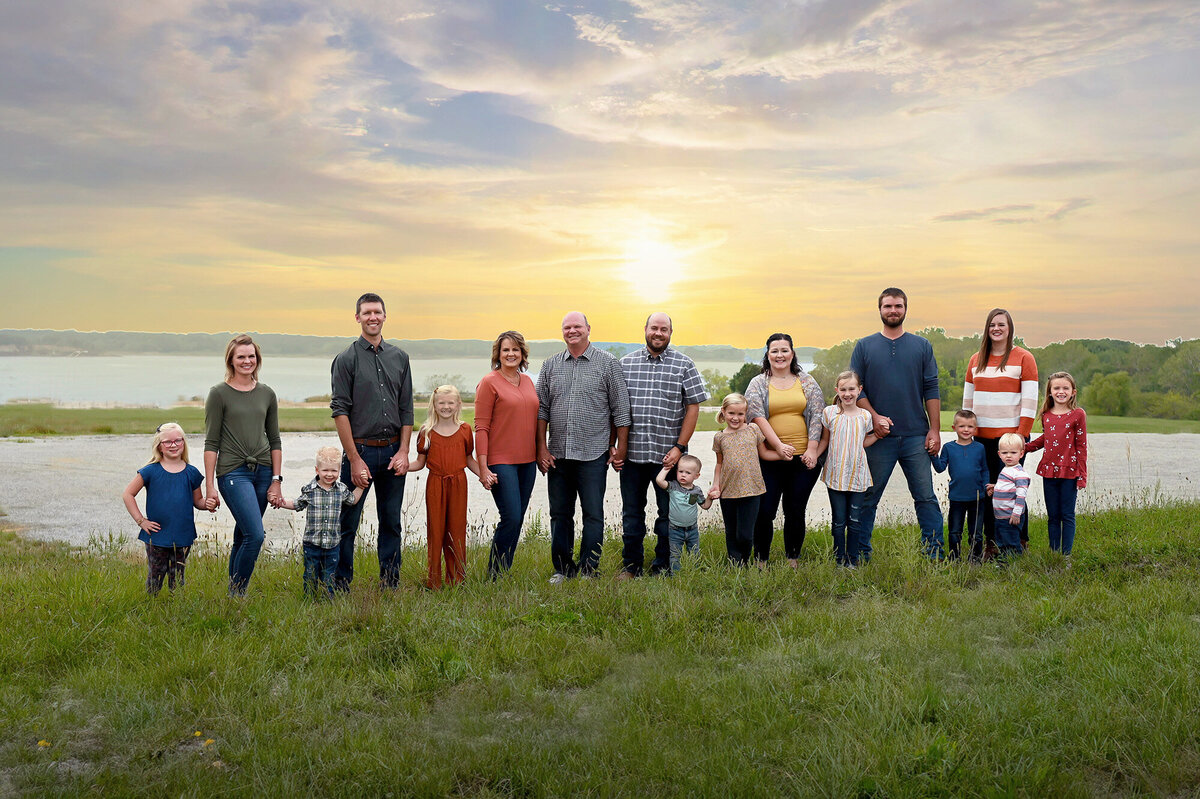 A large family stands next to each other and holds hands in front of a lake for their Pella Family Portraits