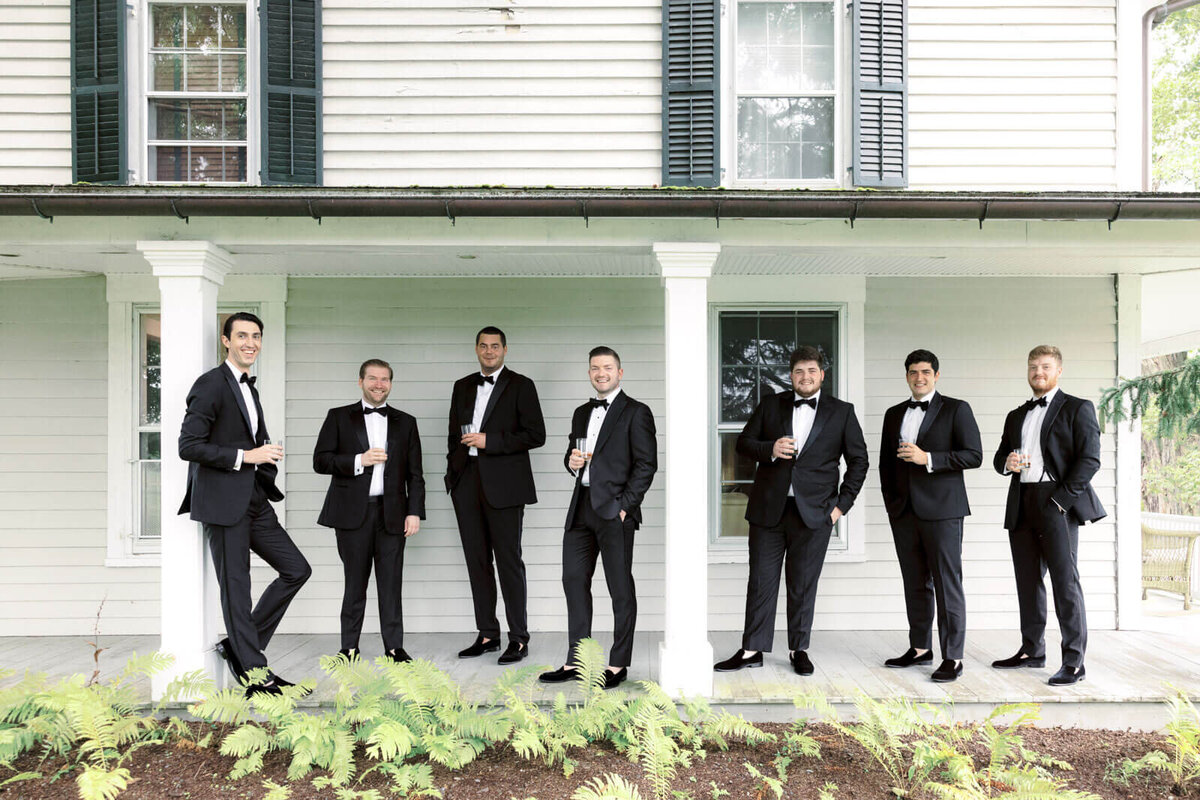 The groom and groomsmen are smiling at the camera, holding a brandy, outside the guest house at Lion Rock Farms, CT. Image by Jenny Fu Studio