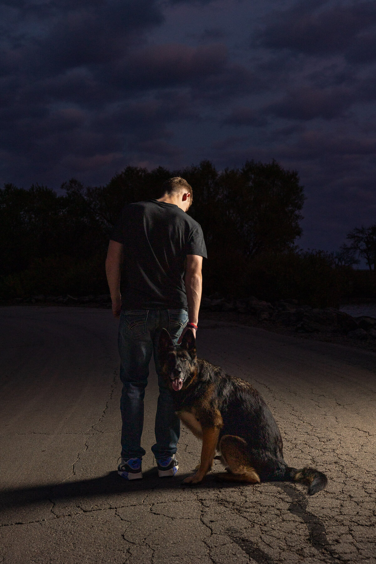 A senior guy facing away from the camera with his dog standing behind him looking at the camera in the dark