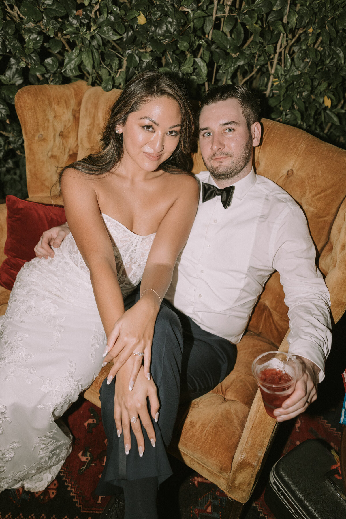 Bride draped over grooms leg posing in vintage style orange chair while he holds a drink