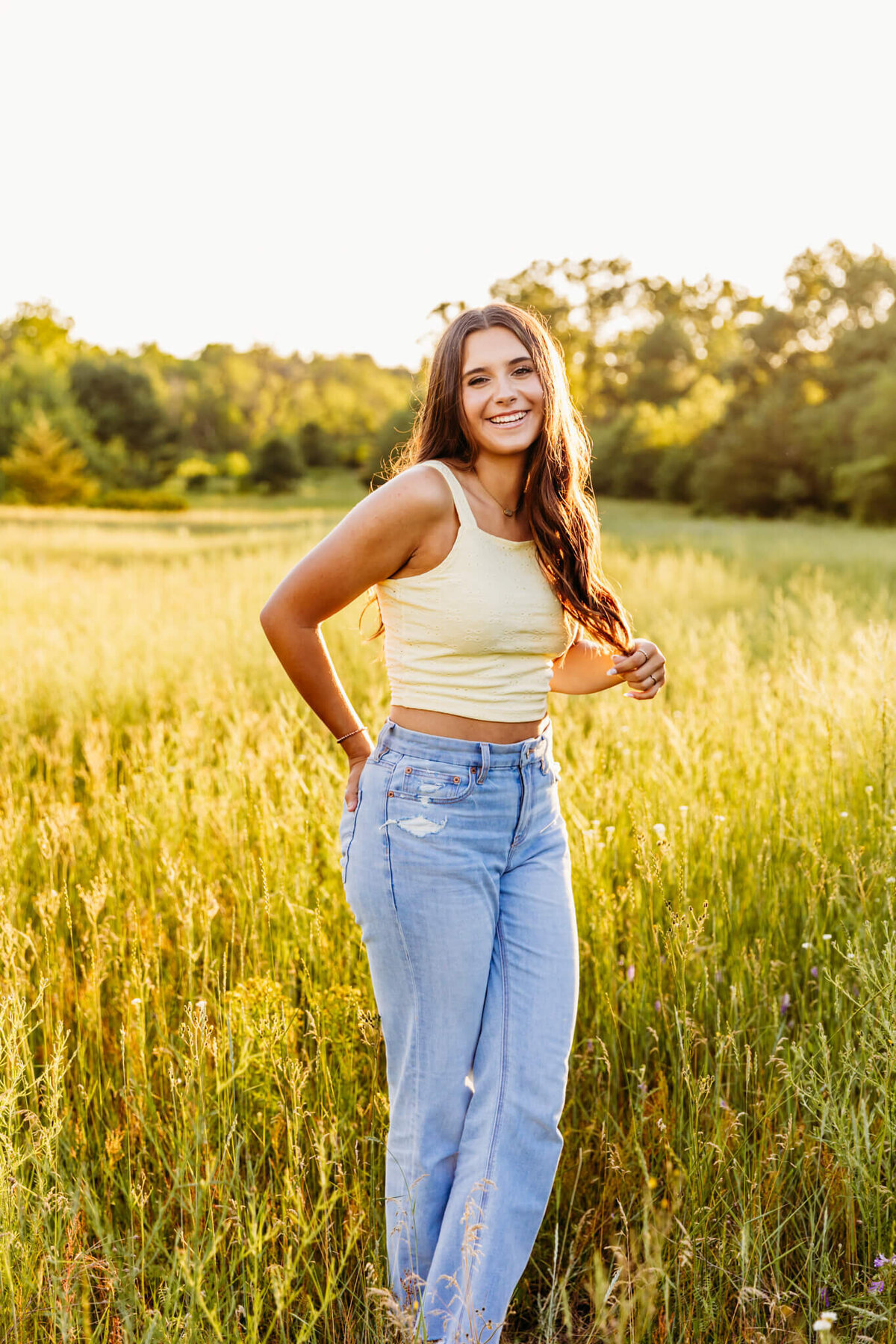 pretty high school girl playing with her brown hair and laughing as she stands in a glowing field captured by Ashley Kalbus Photography