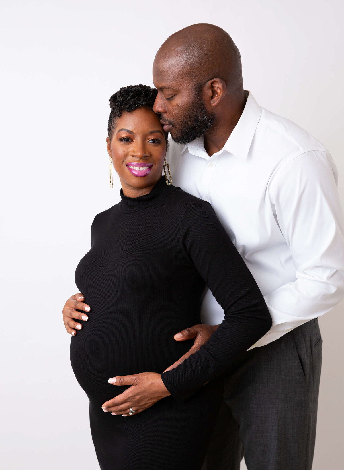 A man leans over kissing his pregnant wife in a black maternity one piece maternity suit after maternity pictures in NYC