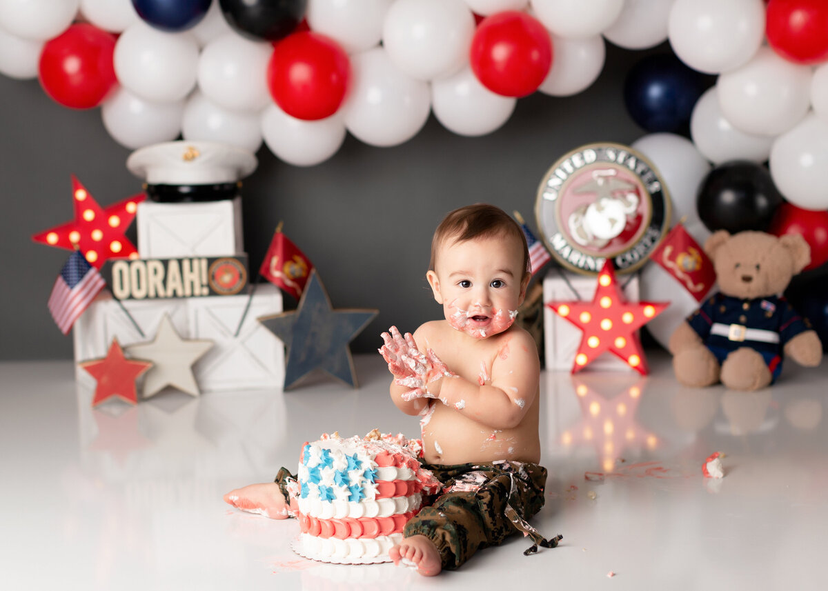 Marine themed cake smash in West Palm Beach and Lake Worth photography studio. Baby boy is wearing camo pants with a red white and blue balloon arch, stars, and american flags in the background The cake is decorated like the US flag. Baby boy has icing on his face, hands, and chest.