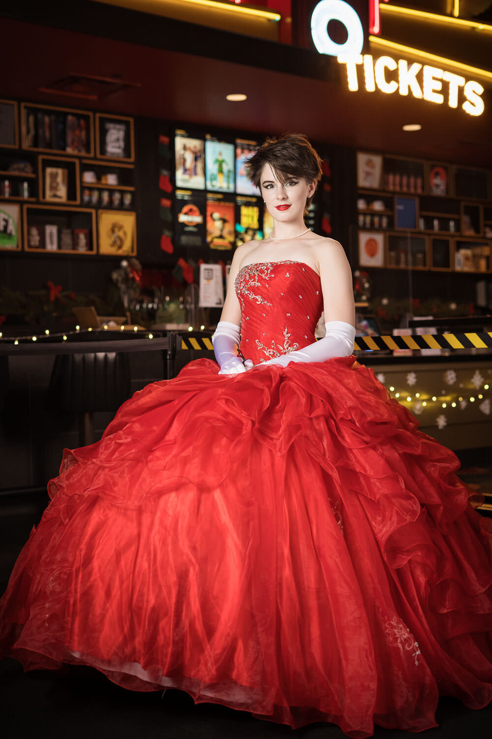 Editorial-style portrait of a glamorous woman in a red formal gown, posing in front of a box office concession stand.