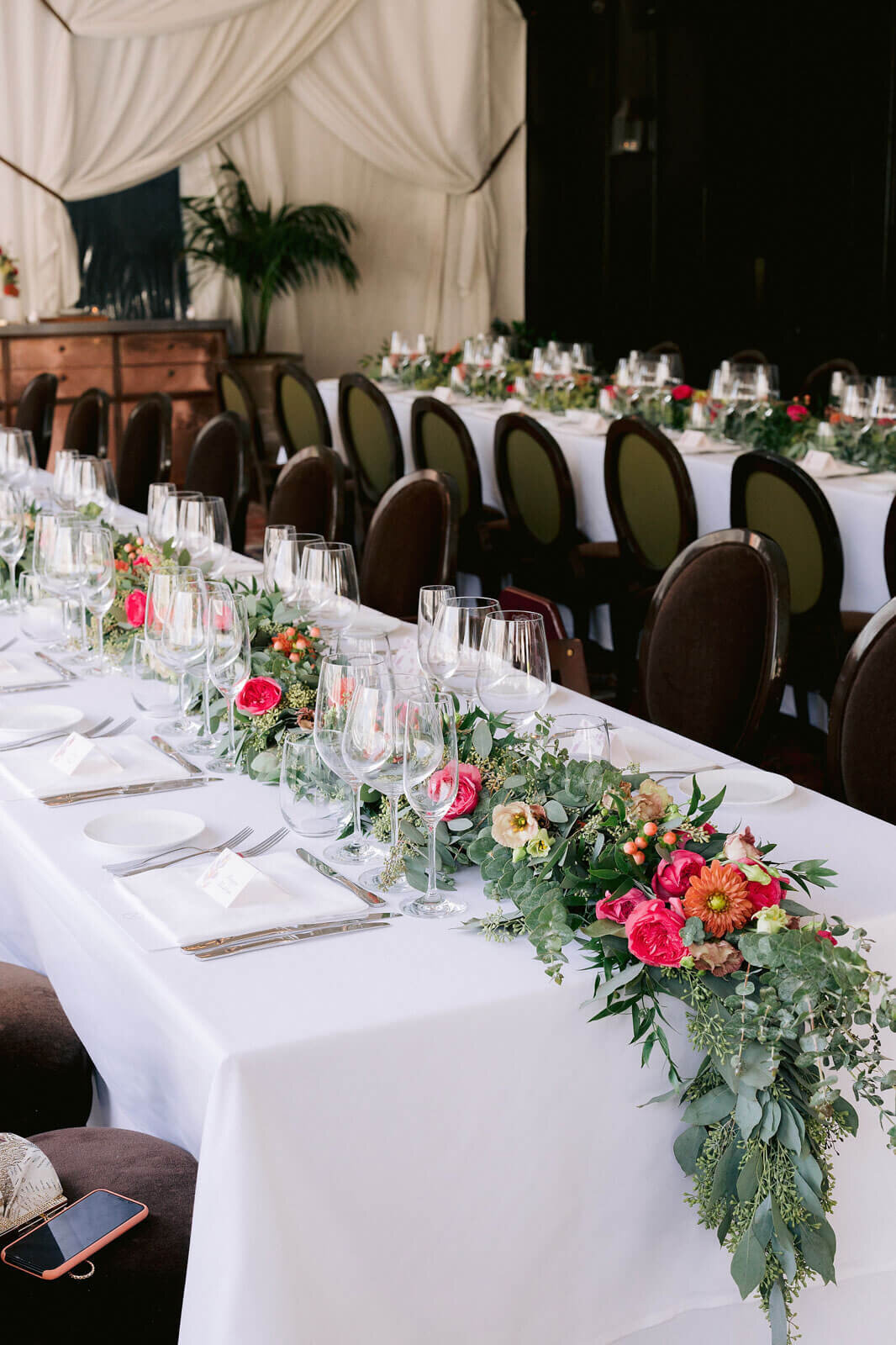 A long wedding dining table with beautiful red, orange, and white flowers and green leaves as the centerpiece. Image by Jenny Fu Studio