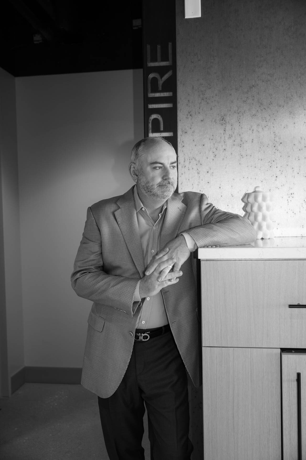 Black-and-white portrait of a contemplative man leaning on a modern cabinet, wearing a light-textured blazer and dress shirt, with 'PIRE' visible in the background, evoking a professional and thoughtful atmosphere.