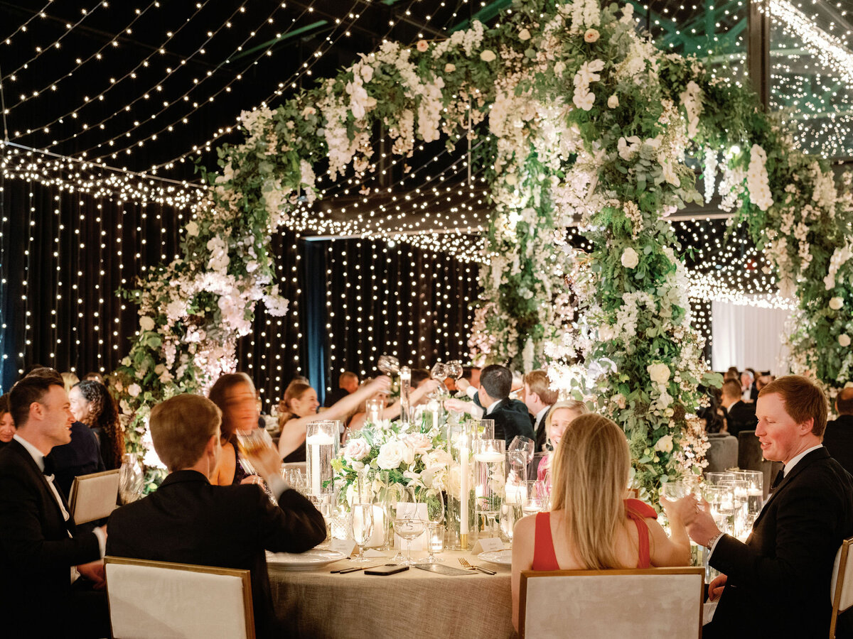Wedding guests raise their glasses in a toast, surrounded by elegant candlelit tables and lush floral decorations.