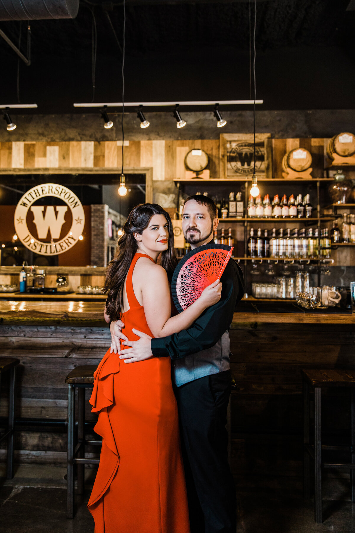 A couple holding each other close in front of a bar in a whiskey distillery during their engagement session in DFW, Texas. The woman on the left is wearing a long, vibrant red dress and is holding a detailed, red, hand fan. The man on right is wearing a black dress shirt, black pants, and a grey vest.