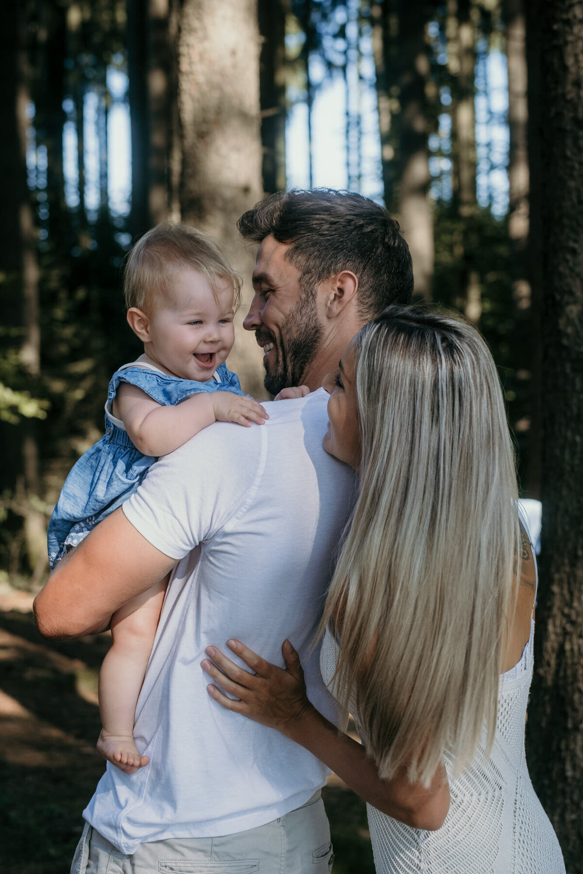 Mom hugs dad, whois holding the laughing babygirl, standing in the soft sunlight of a forest.