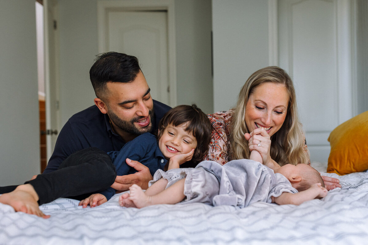mom, dad, and 4-year-old son leaning over newborn on bed while mom holds baby's hand to her face