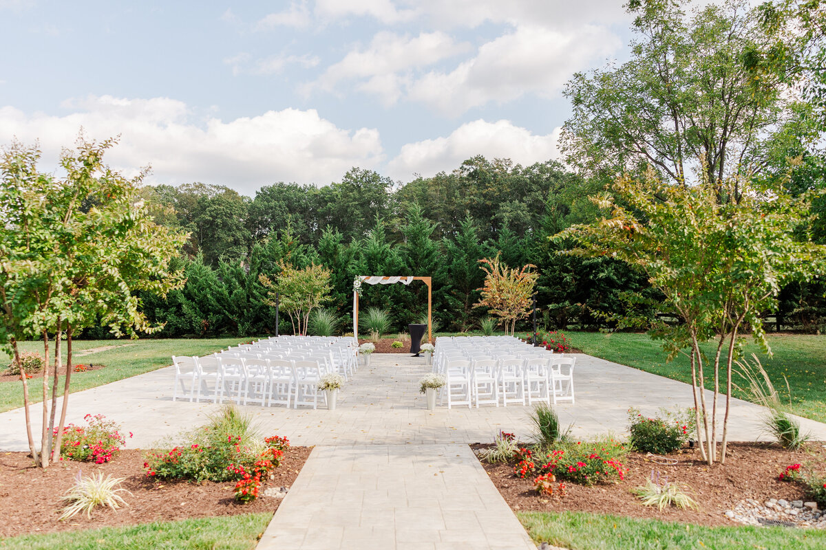 A picturesque Leesburg VA wedding venue features a grassy outdoor ceremony setup with rows of white chairs facing a wooden arch draped in white fabric. Green trees border the area, complemented by small red flowers in the foreground, all under a partly cloudy sky.
