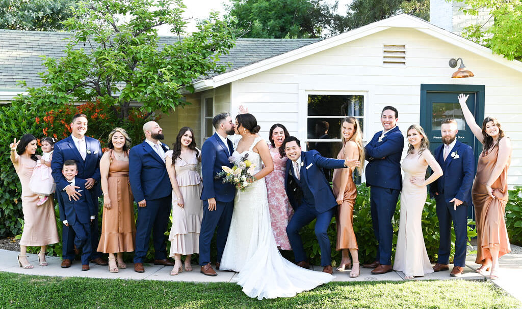 A bride and groom kissing with their wedding parties celebrating behind them.