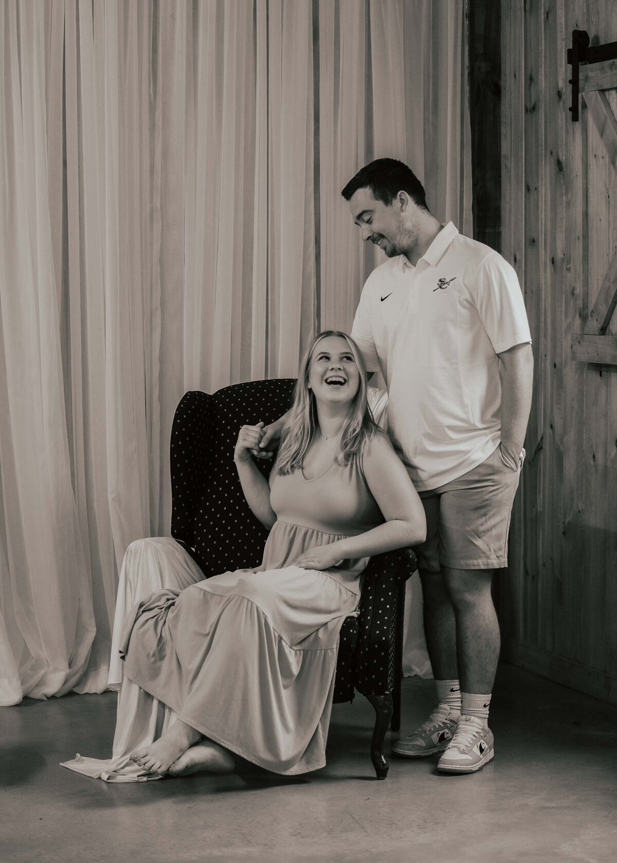 Maddie Rae Photography black and white couple image, the girl is sitting in the chair looking over her shoulder at her fiance who is standing behind her