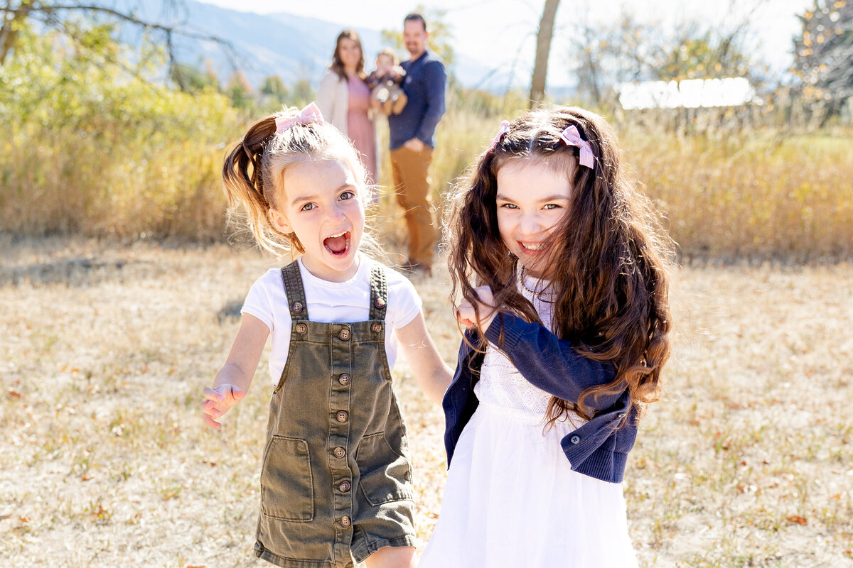 Two little girls with high ponytails and bright smiles run through a sunlit autumn field, their laughter radiating pure joy. Their parents stand in the background, watching them lovingly.