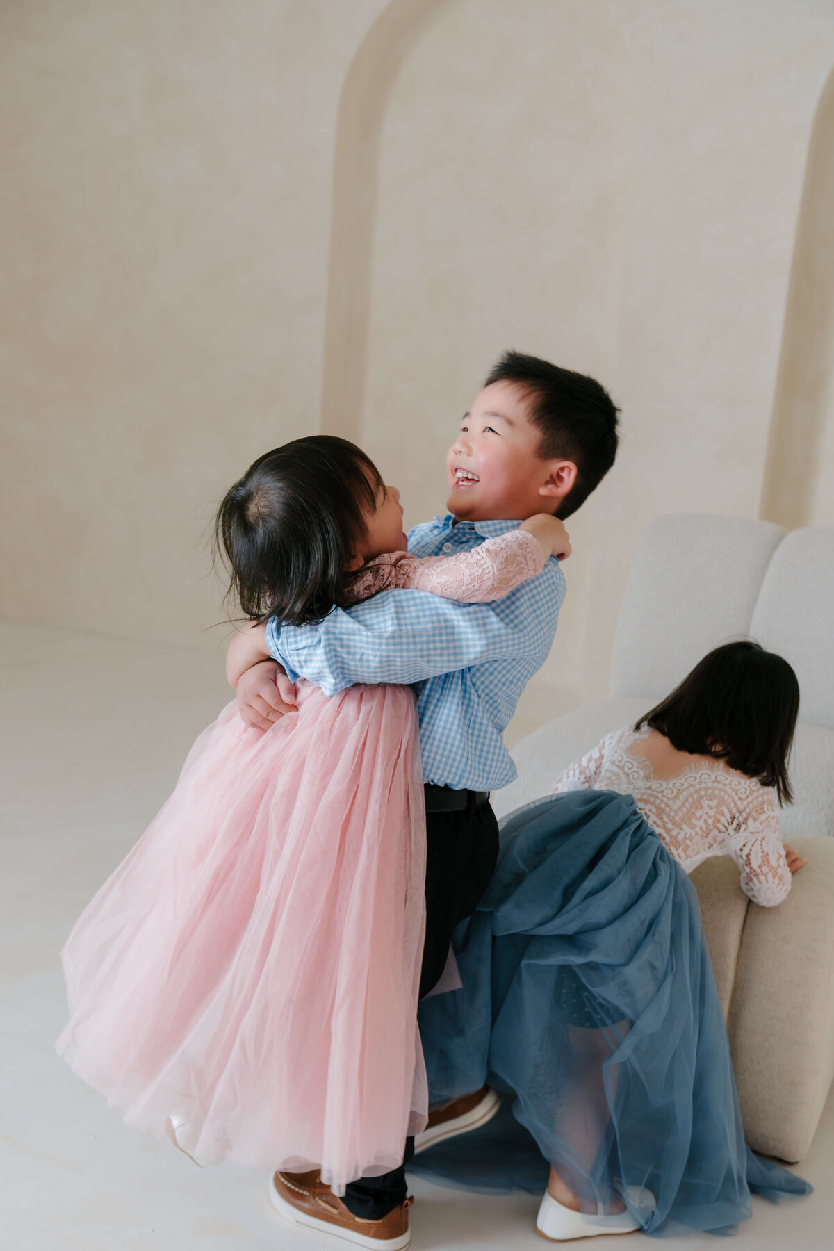 Brother and sister share a joyful hug in a studio with soft neutral tones and minimal backdrop.