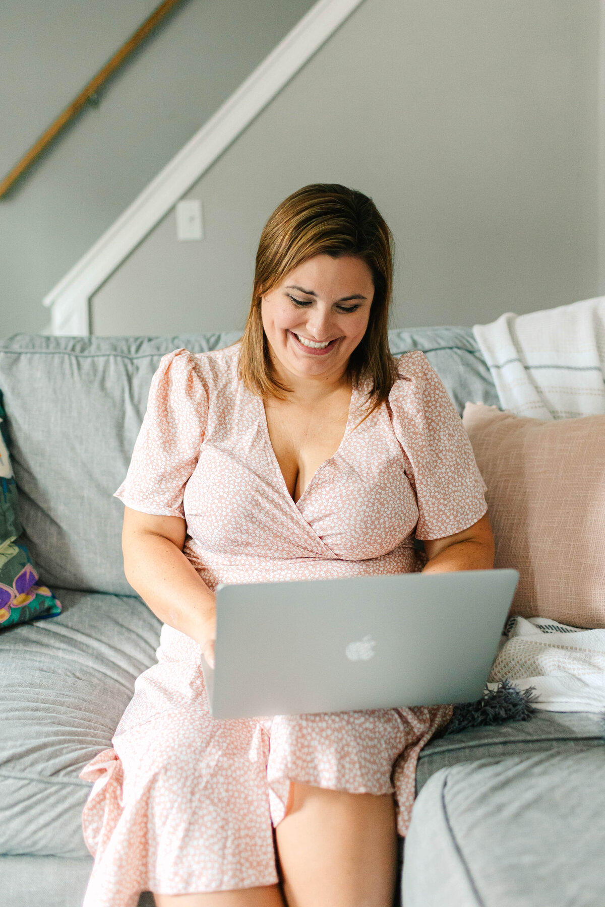 A woman in a pink floral dress sits on a gray couch, smiling as she works on her silver laptop, surrounded by cozy pillows.