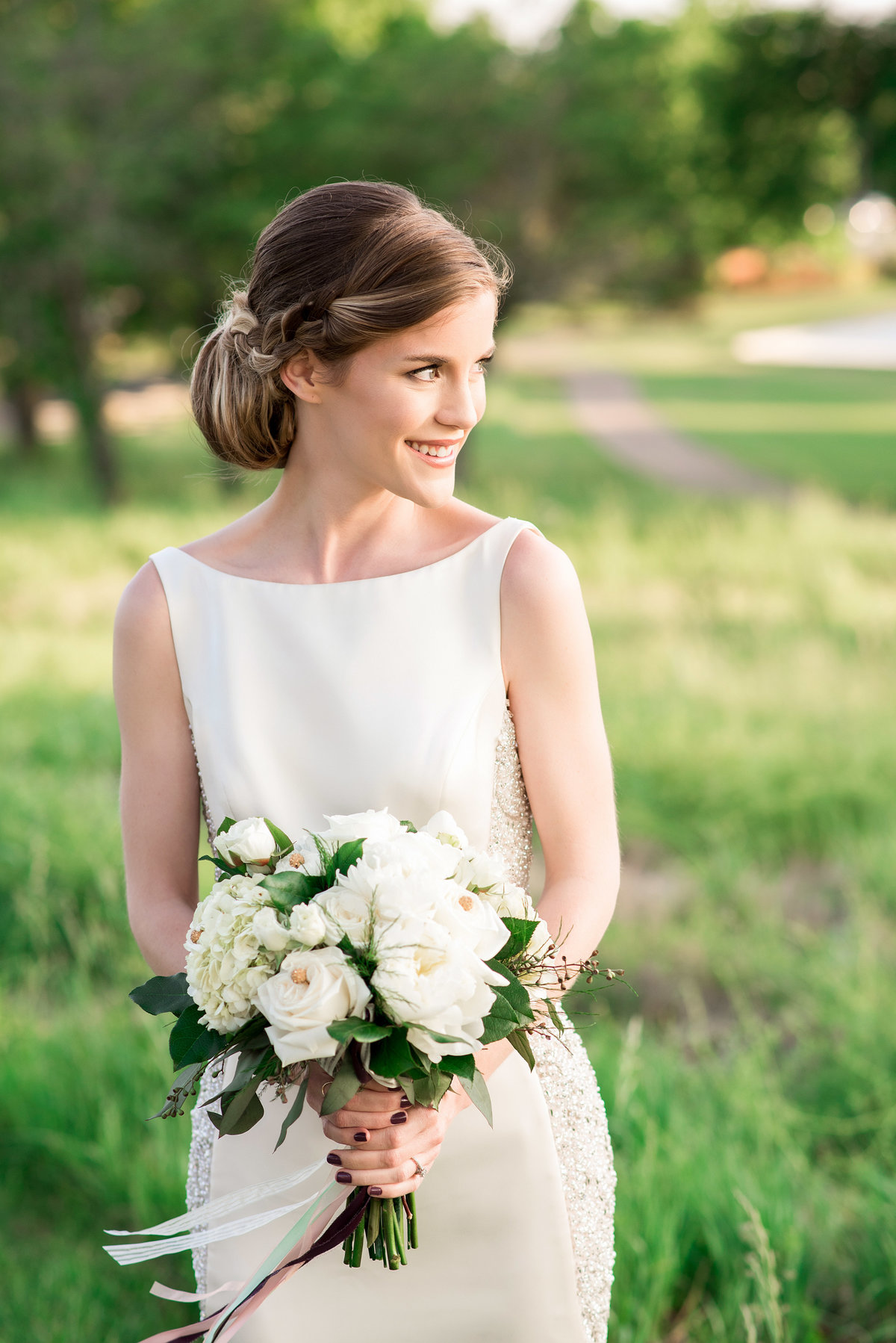 The bride stands in an overgrown green field looking off to the right. Her hair is in a low bun and she is wearing a satin sheath wedding dress with a sleeveless bateau neckline. She is holding a large round bouquet of white flowers with long white ribbons that flutter in the breeze.