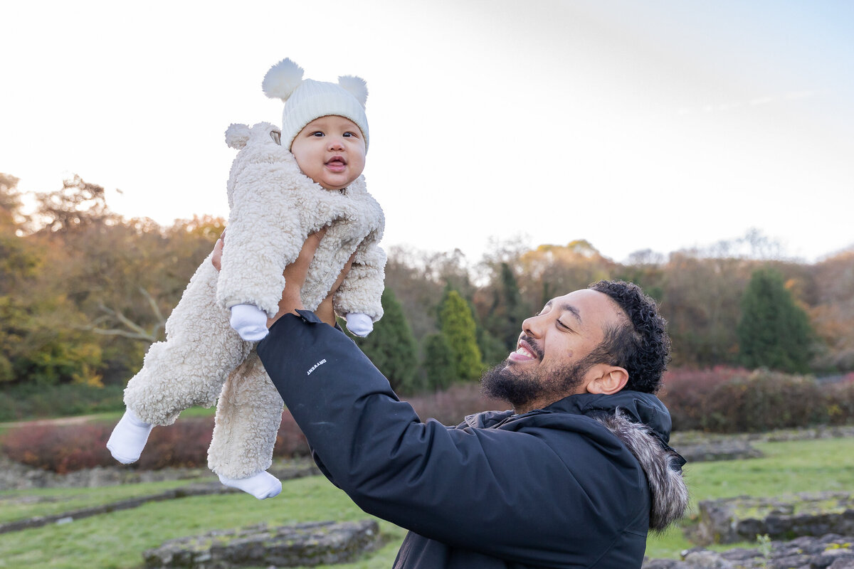 A joyful father lifts his baby, who is wearing a fluffy bear onesie, into the air in a scenic park during autumn.