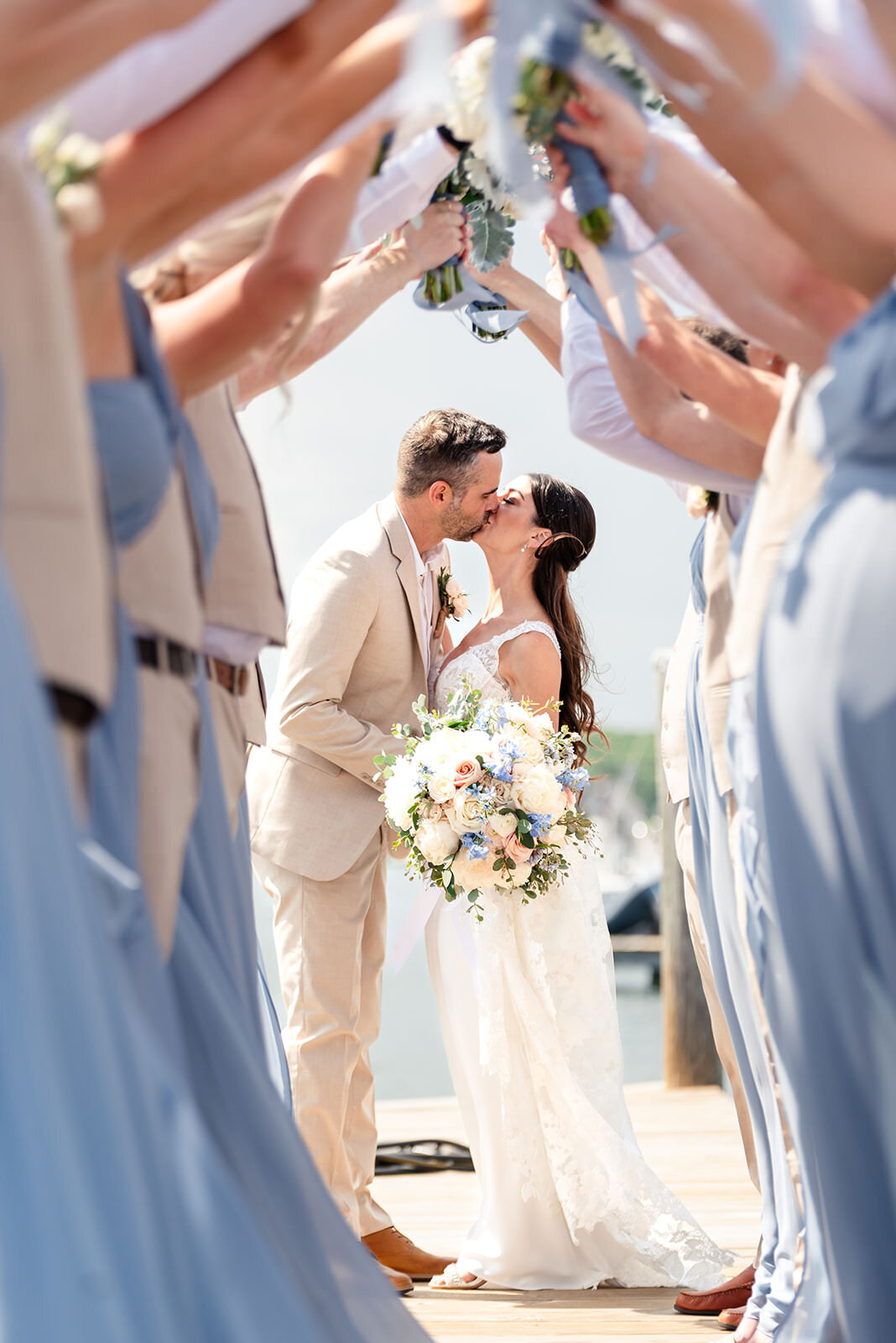 The bride and groom share a kiss under an archway made by the wedding party holding up bouquets, on a dock with a clear sky above.