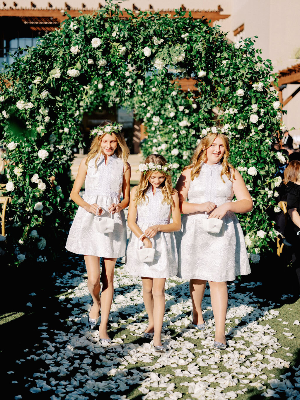 Adorable flower girls in white lace dresses and floral crowns walk down an aisle covered in rose petals, adding a magical touch to this wedding ceremony.