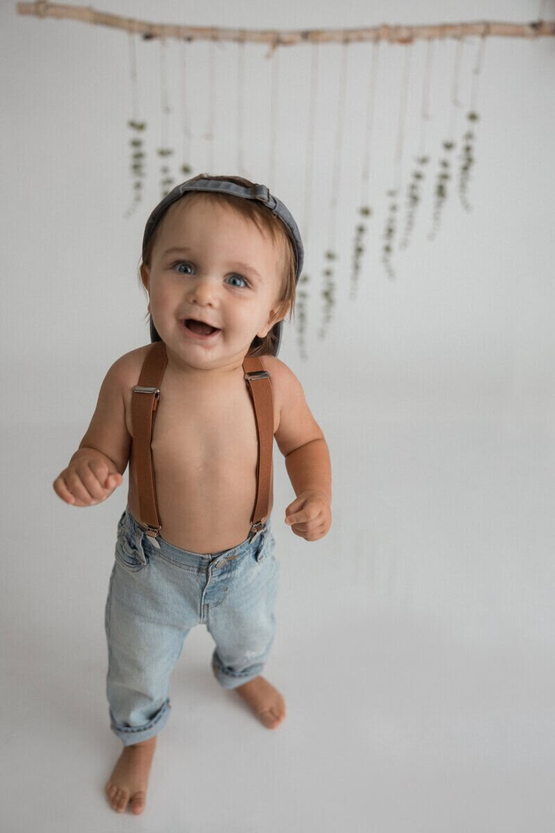 Smiling baby boy wearing denim jeans with suspenders and a cap, standing in front of a minimalistic background with hanging greenery.