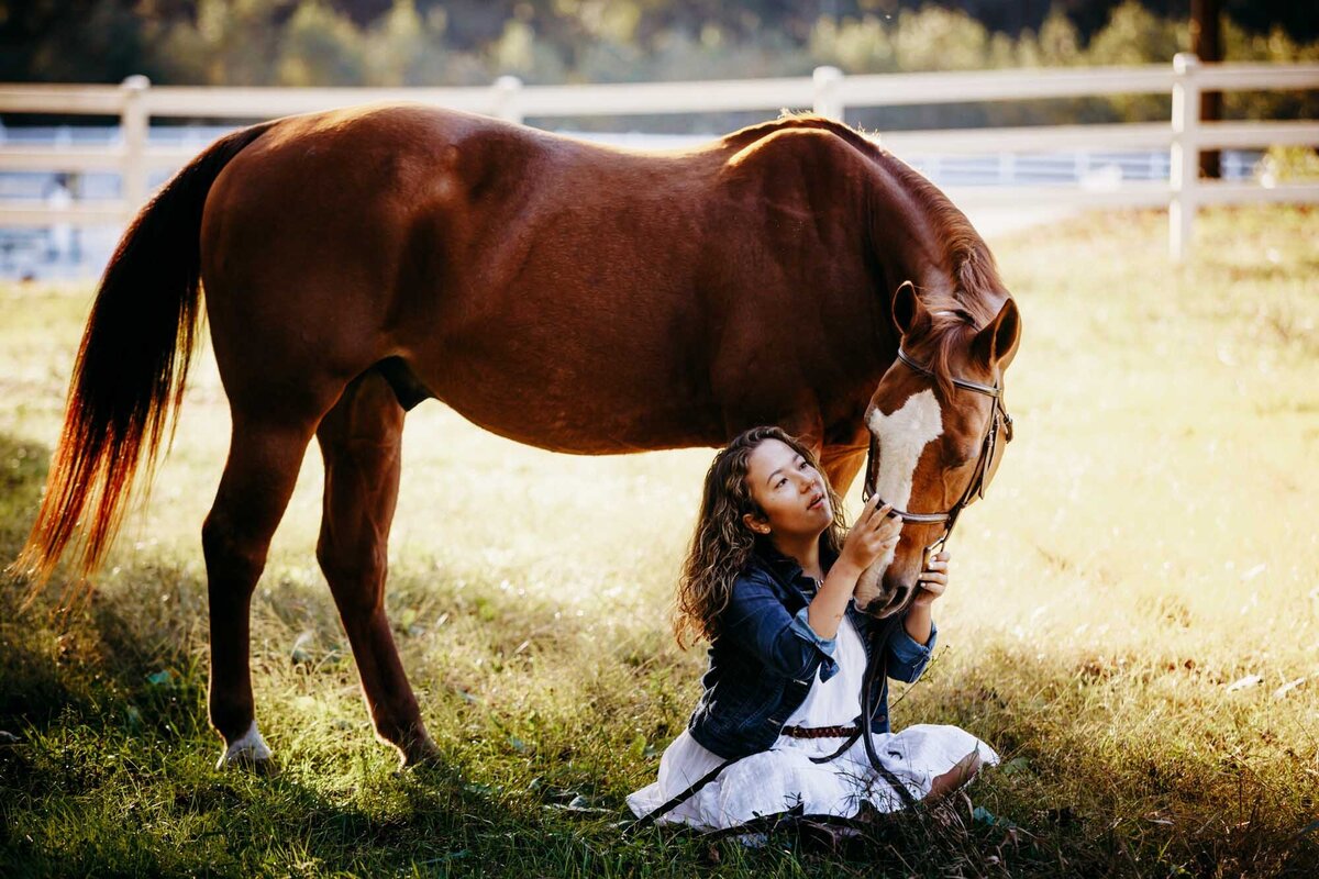 Senior portrait of a girl sitting on the ground while her horse looks down at her.