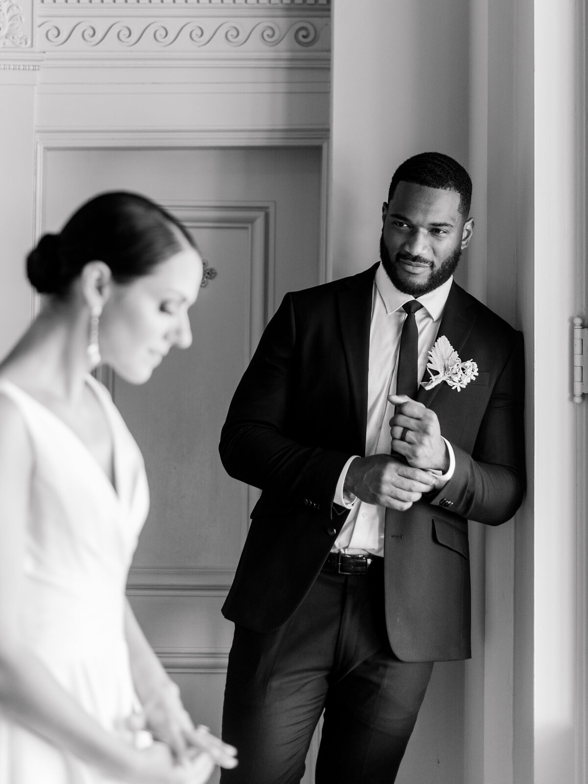 Black and white editorial wedding portrait at Cairnwood Estate captured by Philadelphia Fine Art Wedding Photographer Elizabeth Kane, showcasing a groom in a tailored black suit watching his bride in an elegant gown with soft, romantic lighting—highlighting timeless East Coast wedding style and refined emotion.