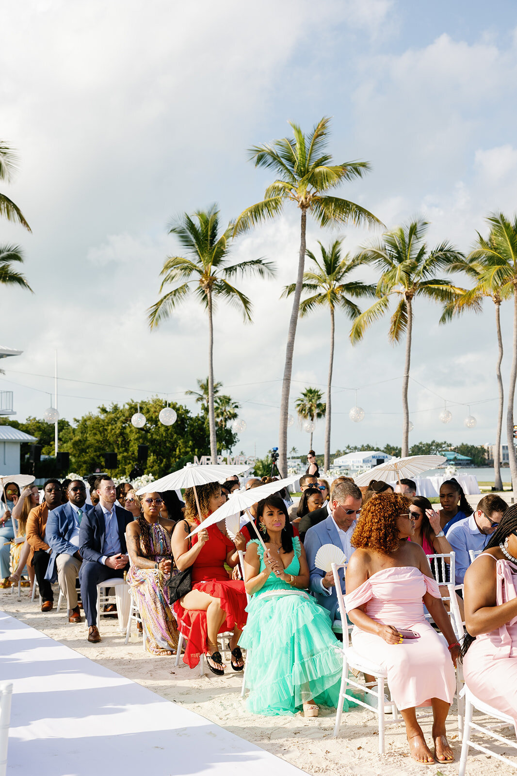 Guests dressed in vibrant colors sit along the white aisle, shaded by elegant parasols, awaiting the bride's entrance at Table Key, Islamorada. Photography by Claudia Amalia Wedding Photography.