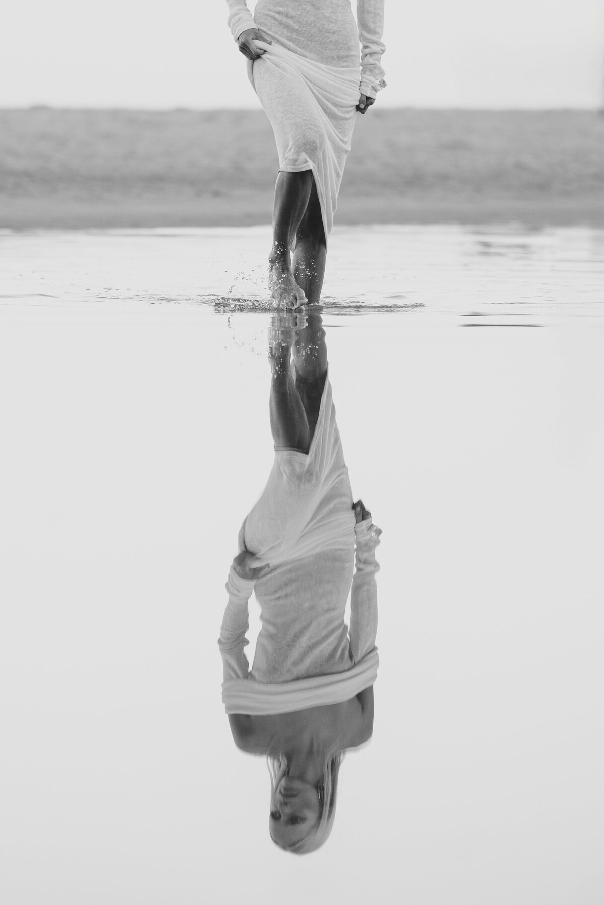 “Zwart-wit foto van model Johanna in een witte jurk die door het ondiepe water loopt in Kaapstad, met haar reflectie zichtbaar in het spiegelgladde oppervlak.
