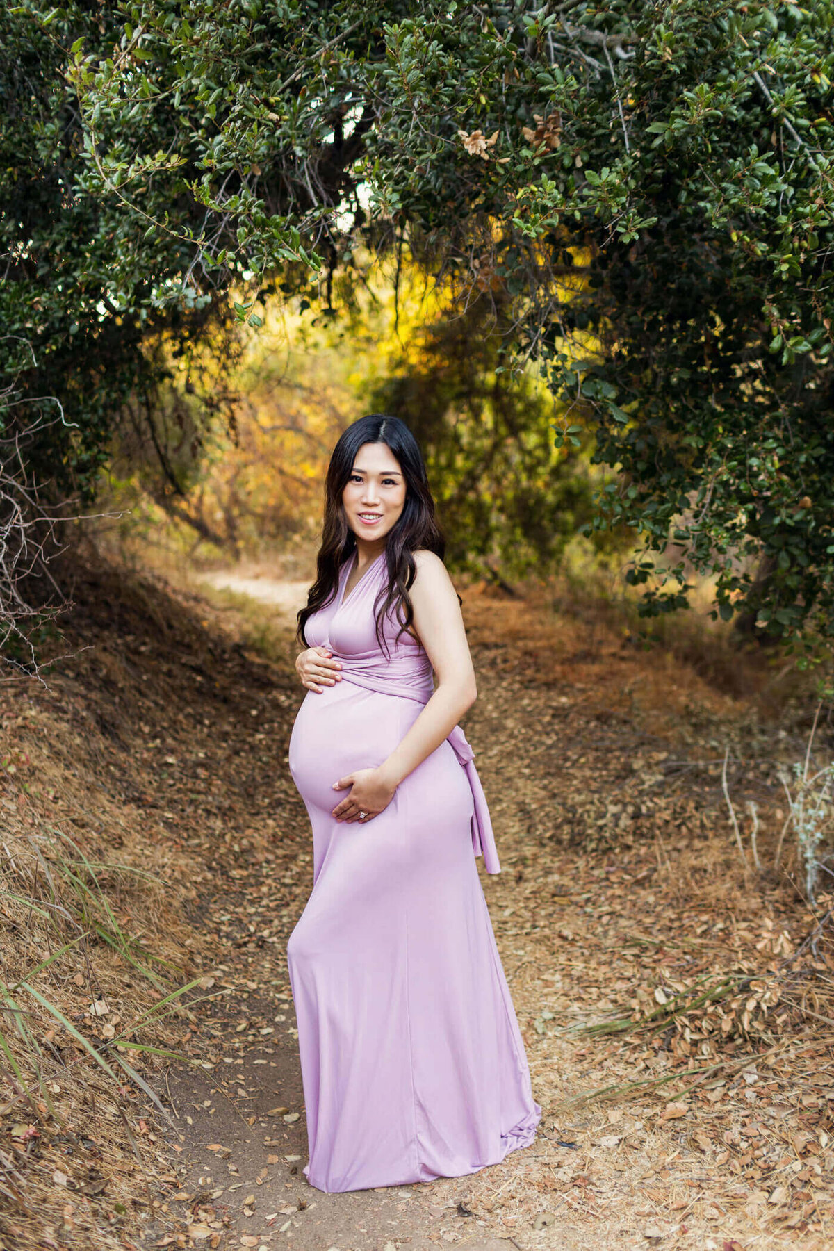 A pregnant woman stands on a wooded path surrounded by lush green trees and foliage. She is wearing a form-fitting, sleeveless lavender dress that highlights her baby bump. Her long, dark hair cascades over her shoulders as she smiles gently at the camera. The natural setting, with its arch of leaves above her, creates a serene and picturesque atmosphere.