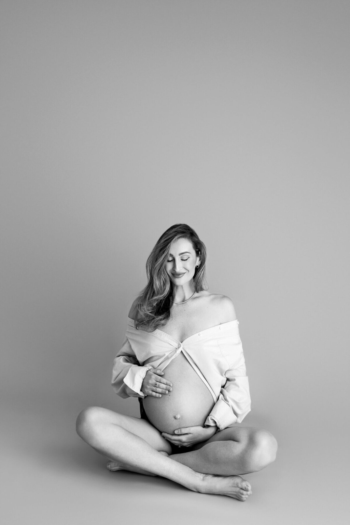 A black and white maternity portrait of a serene pregnant woman sitting cross-legged on the floor, wearing an unbuttoned oversized shirt that reveals her baby bump. She gently cradles her bump with both hands, one hand resting above and the other below, while she gazes down with a peaceful, contented smile. The simplicity of the background and her relaxed posture create an intimate and timeless image, capturing the beauty and tranquility of pregnancy.