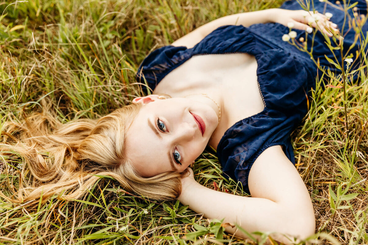 stunning senior girl in a blue dress laying down in a flower field with pink lips captured by Green Bay family photographer Ashley Kalbus