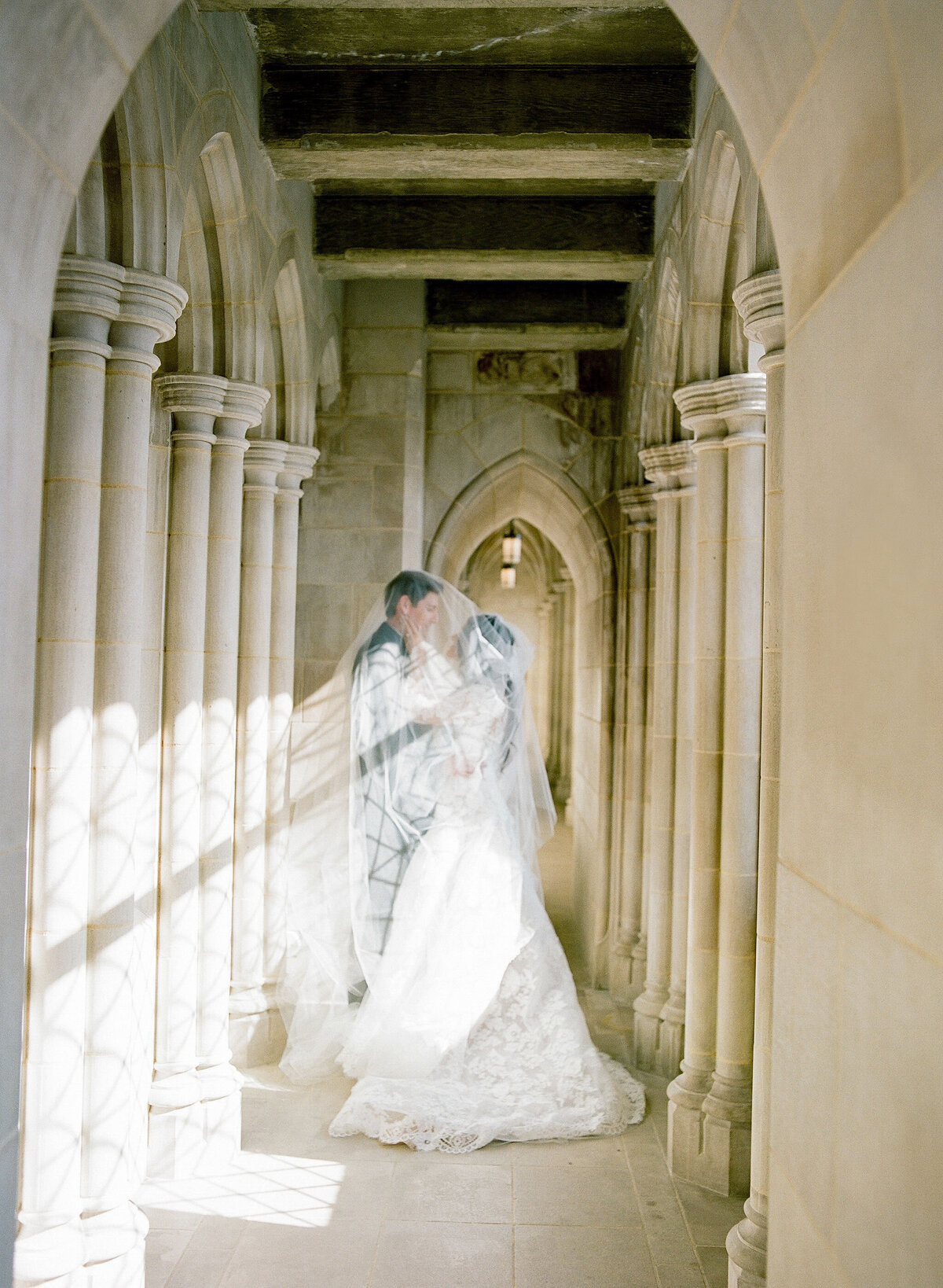 This photo was taken at Washington D.C., Washington National Cathedral by photographer Twah Dougherty. A couple stands in a narrow, sunlit stone corridor adorned with arches and columns, sharing a kiss beneath a delicate veil. The bride is dressed in a white gown, and the groom's dark suit is faintly visible under the veil. The scene is ethereal and romantic. The photographers responsible for capturing this beautiful moment are Current Films as Videographers.
