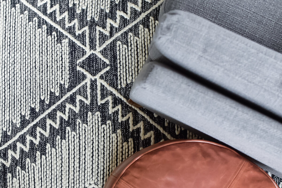 A grey and black patterned rug with a brown leather ottoman and grey armchair