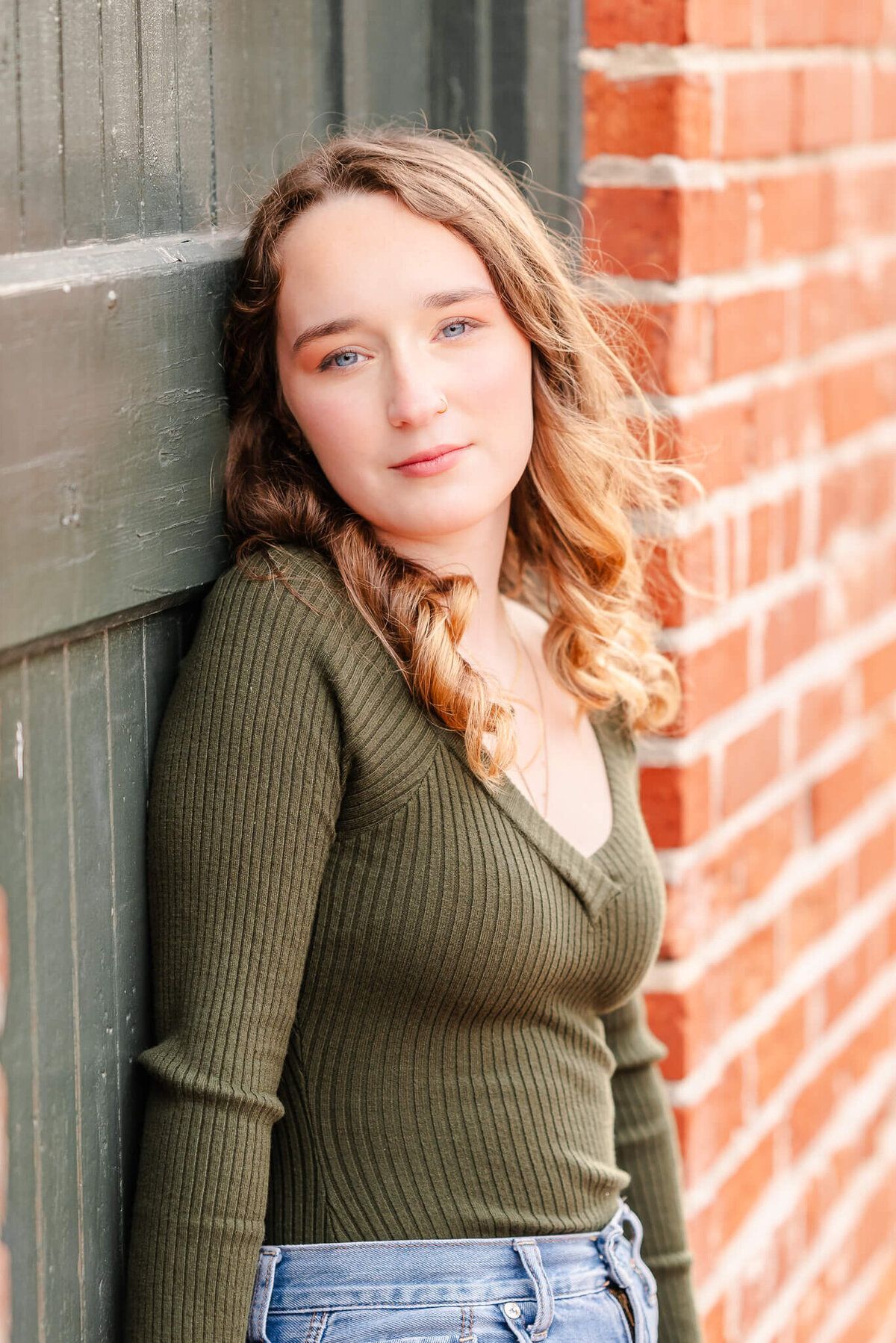 A high school senior, wearing jeans and a green sweater, leans against an old painted door in a brick building.