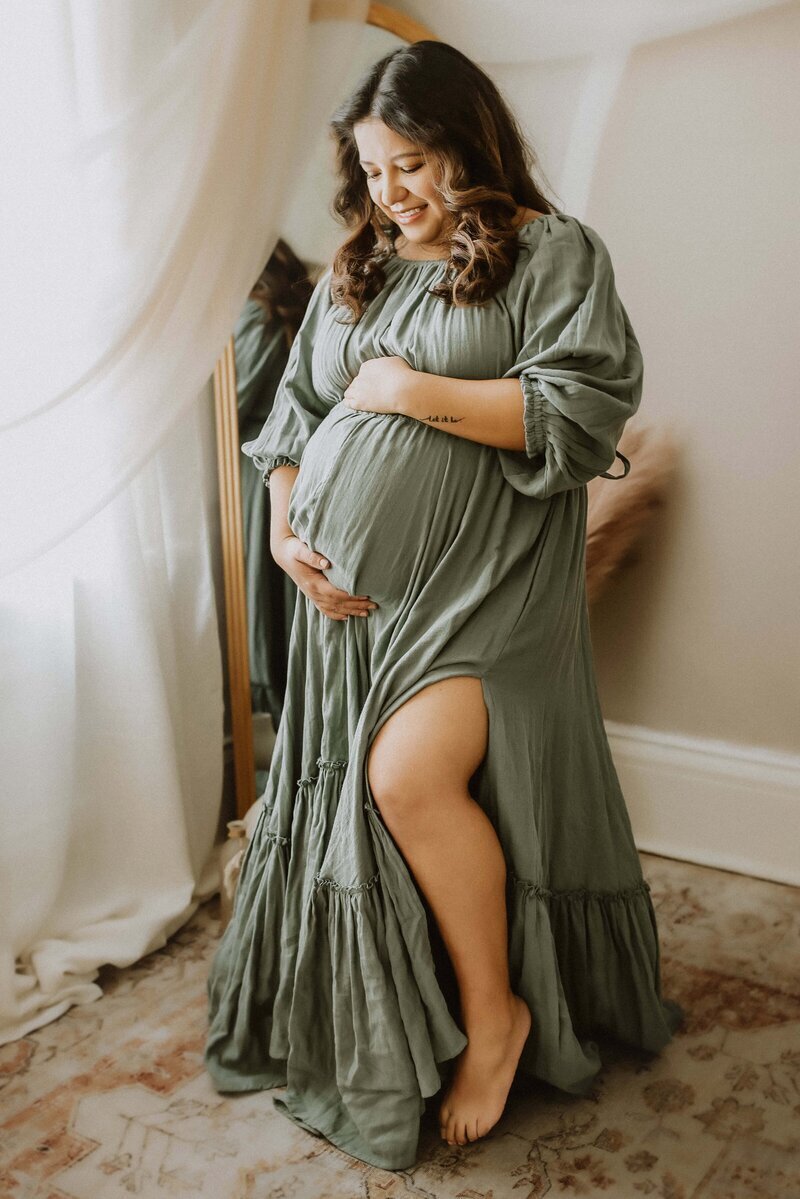 Pregnant woman in a green dress gently holds her belly, standing barefoot in front of a mirror beside flowing white curtains.