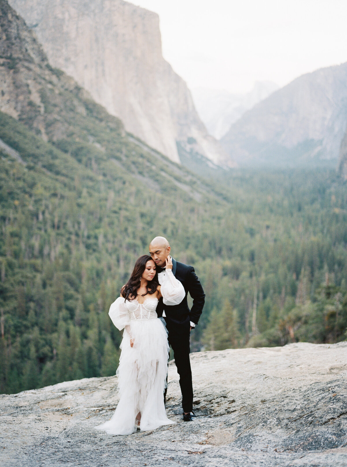 bride and groom standing on a cliff ledge with mountains in the background. The bride has her back to the groom with her left hand caressing his face taken by Mansfield Wedding Photographer, Annie Trammel