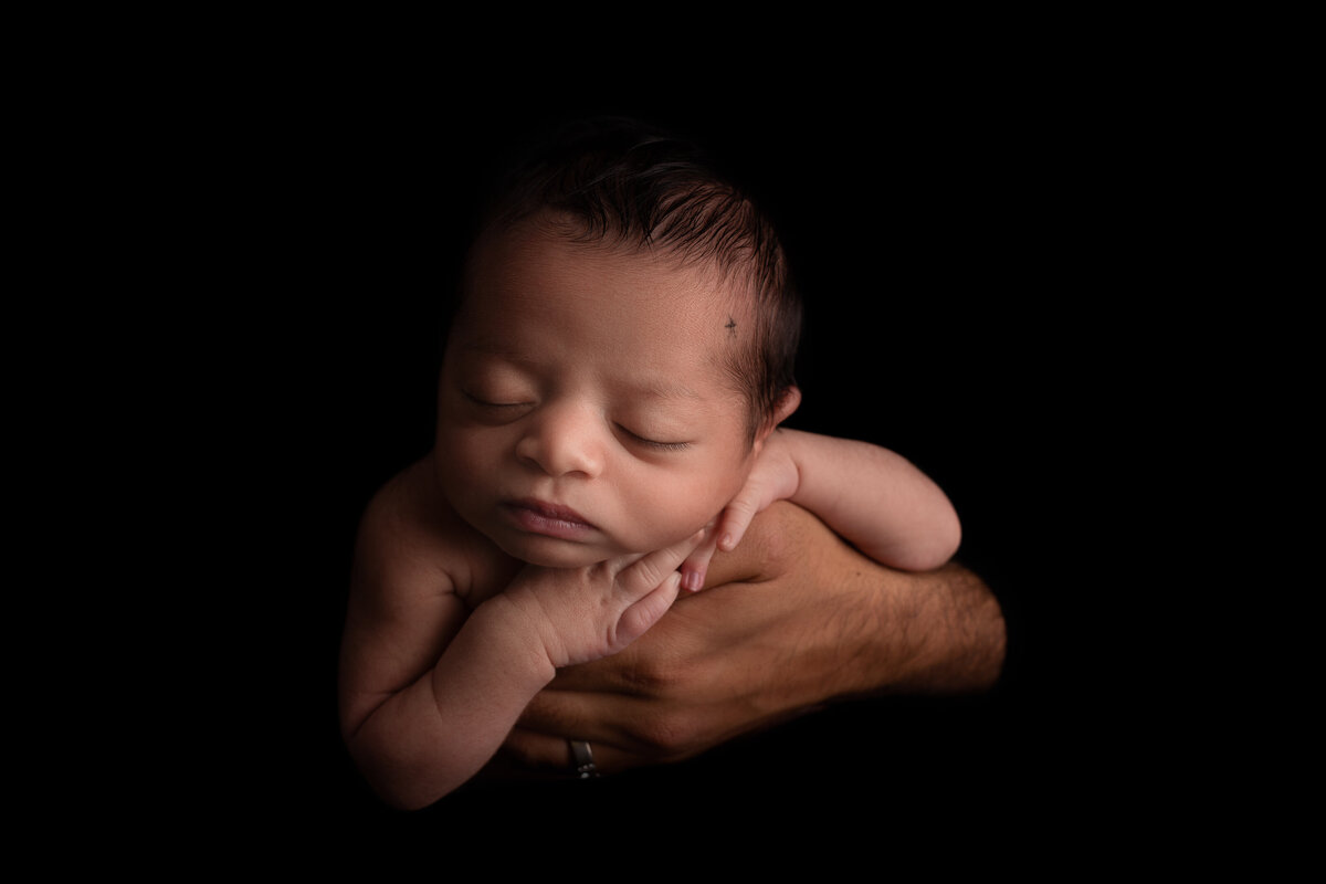 Newborn baby sleeping on his dad's hand on a black background.