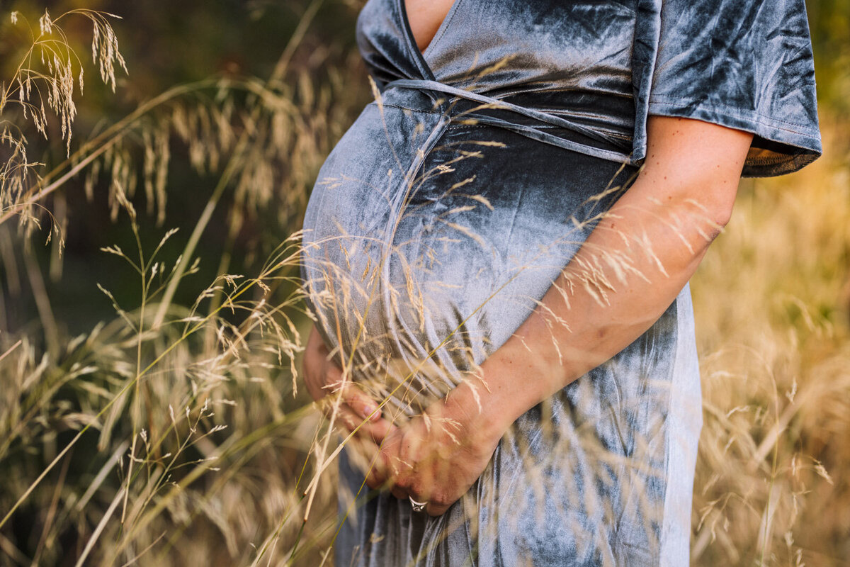 pregnant mom with blue dress standing in tall yellow grass holding her baby bump