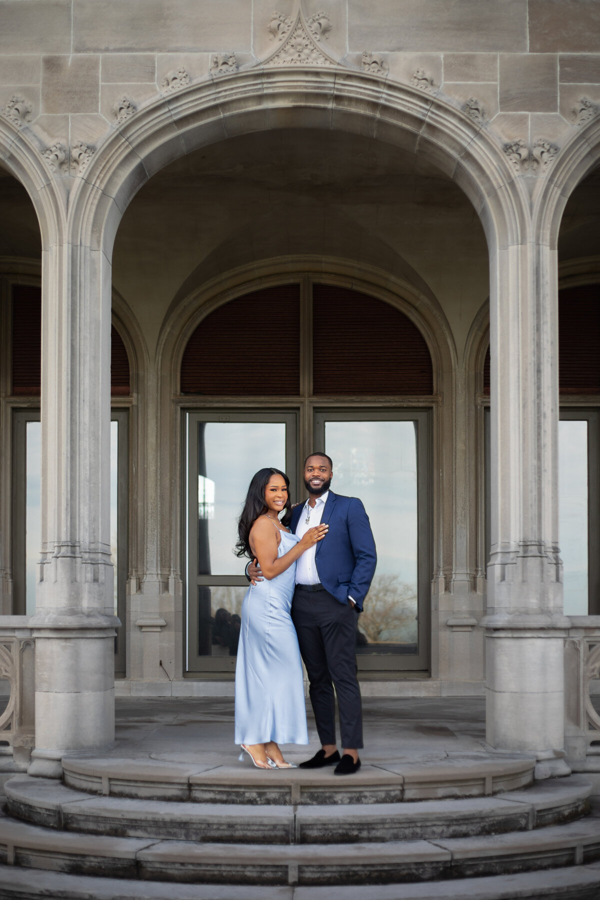 Ochre Court Mansion  | Kelsey Sheehan Photography Timeless Rhode Island Weddings | A couple stands closely under an elegant stone archway. The woman in a light blue dress smiles, holding the man in a navy suit, conveying warmth and joy.