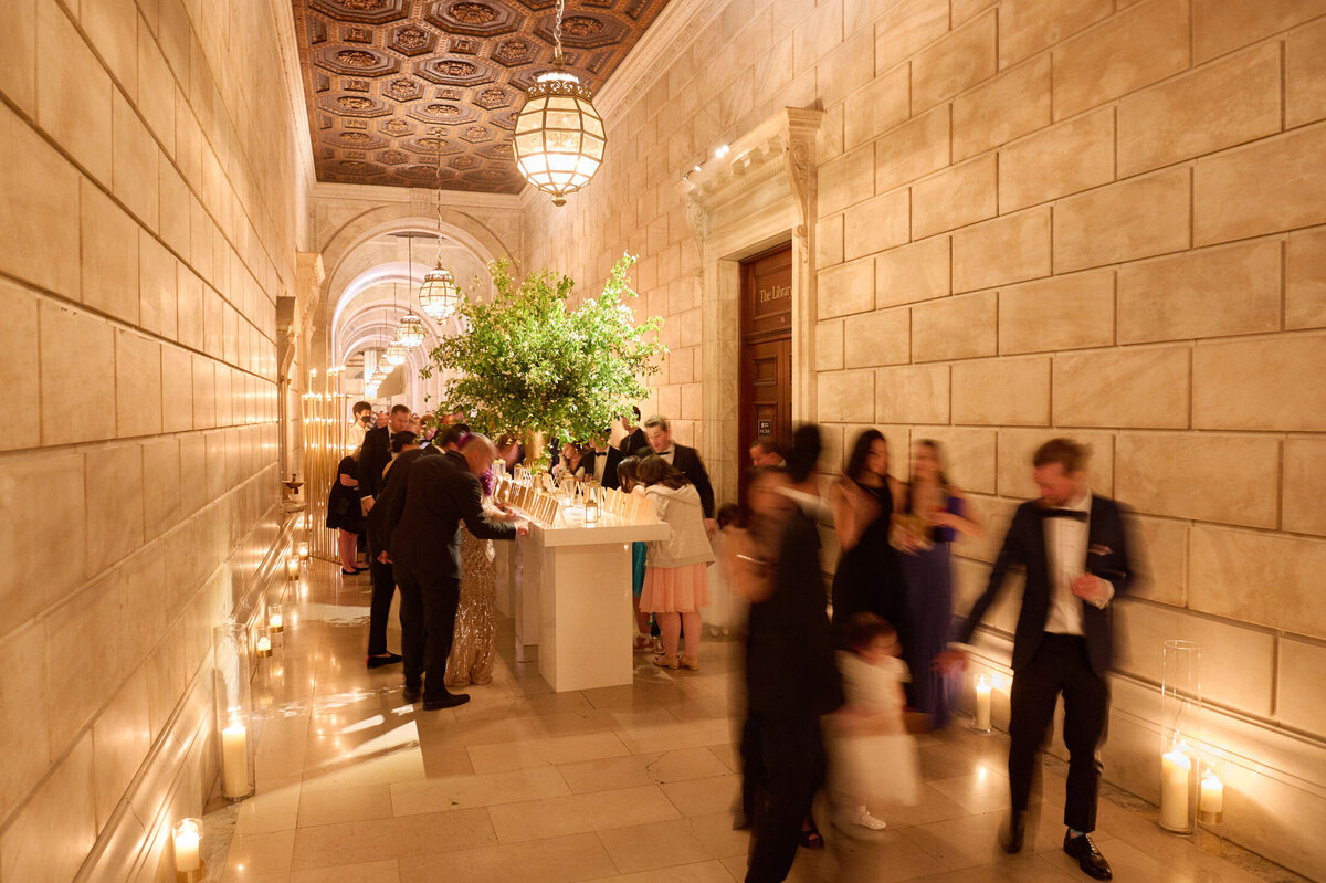 The photo was taken at The New York Public Library in New York City by photographer Shawn Connell. A formal event is happening in an elegant, ornately lit hallway with stone walls and a high ceiling adorned with intricate designs. Guests in formal attire mingle and converse near a bar set up with drinks, while a few people walk through the hallway. Candles line the floor edges. Videographers from Aaron Navak Films were also present to capture the event.