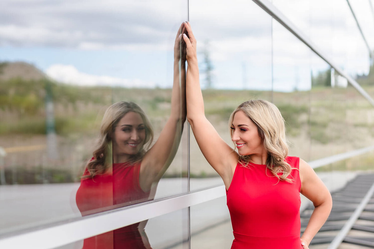 Anita Parker wearing a red dress and leaning on a glass wall, looking at her reflection outside the Kelowna Aviation Museum.