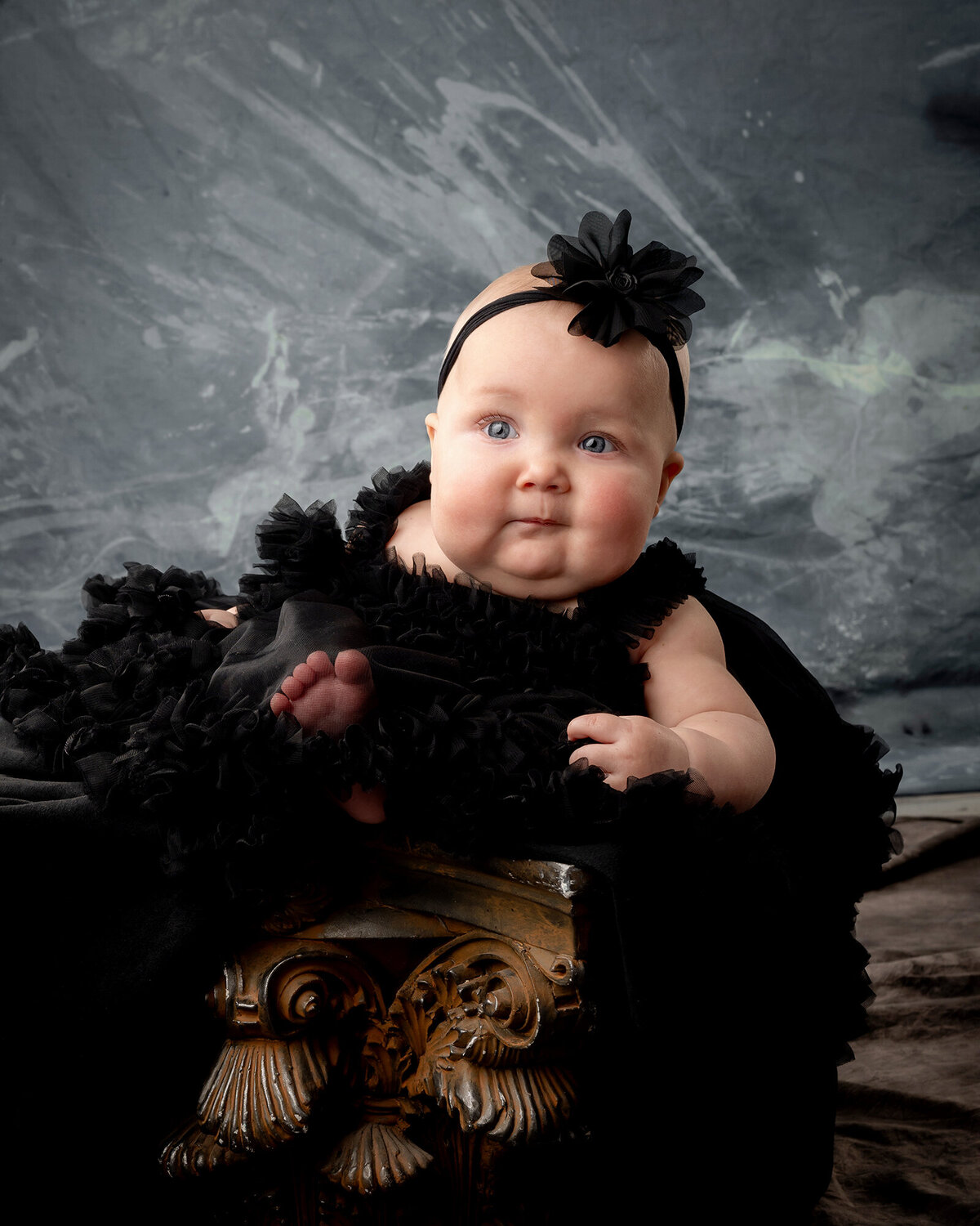 A baby girl sits in an ornate box in a black dress