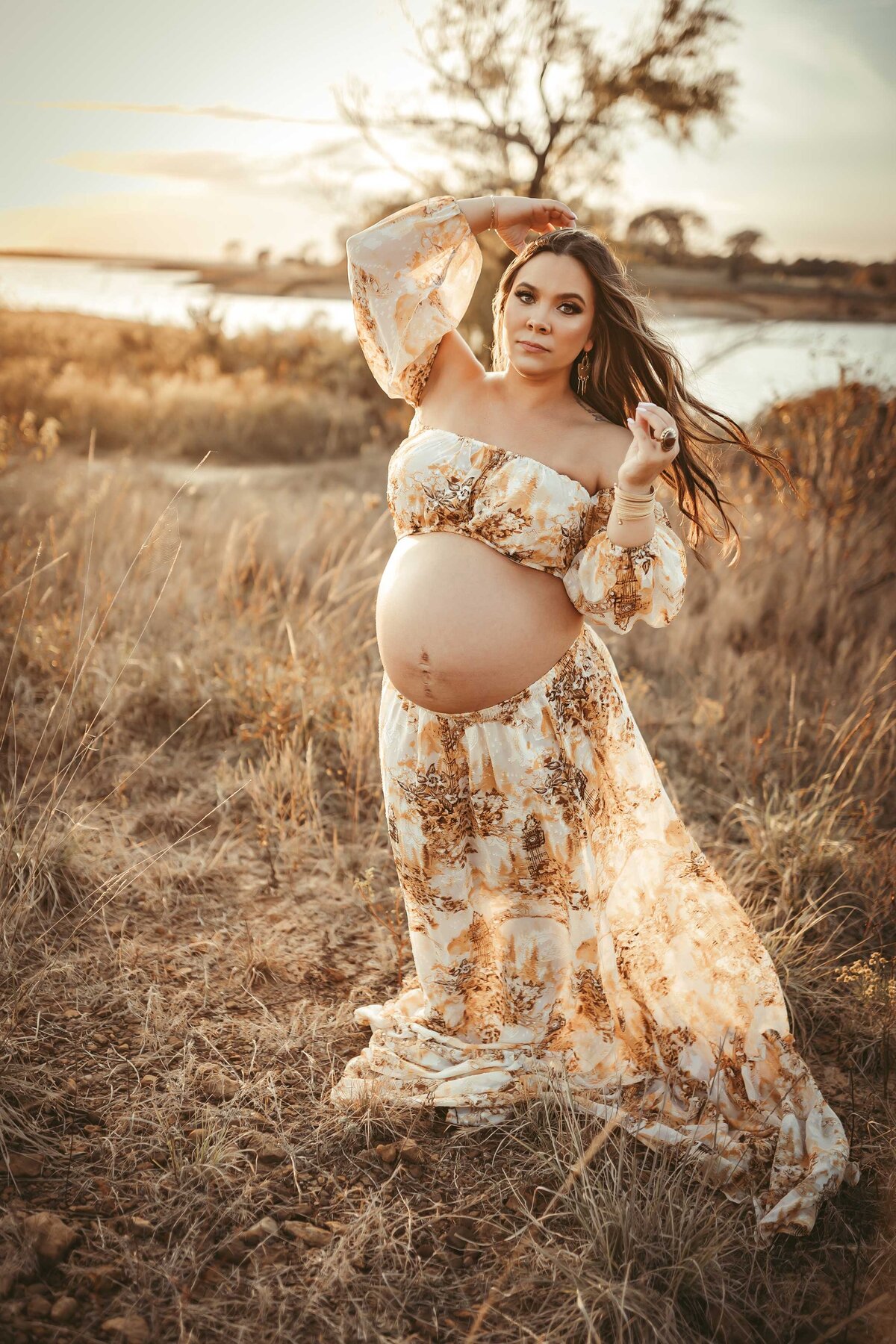 Woman in a Dallas maternity session in a field of grass. Her dress and hair are moving with the wind. The sun is shining and reflecting on the water.