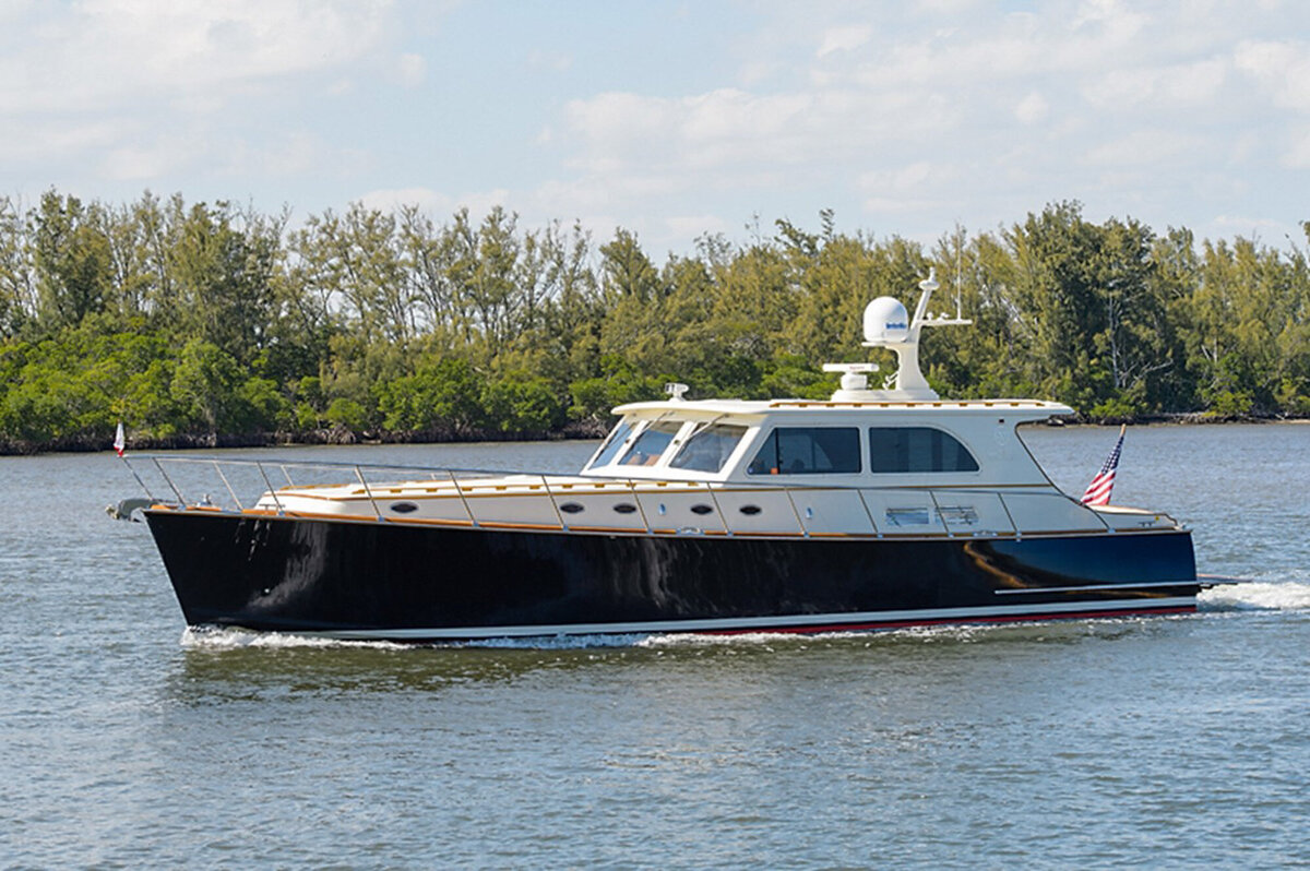 A black and white luxury motor yacht cruises on a calm body of water with green trees lining the background. An American flag is visible at the stern of the boat under a partly cloudy sky.