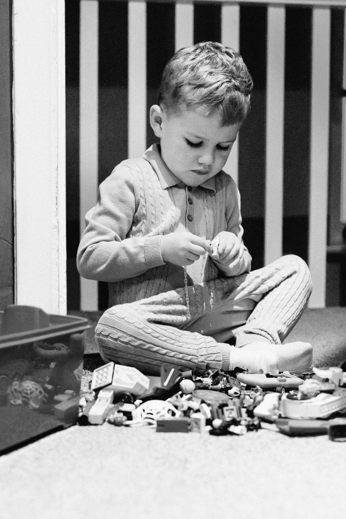 A young boy plays with toy cars on the floor, intently threading a string through them in a black and white photo.