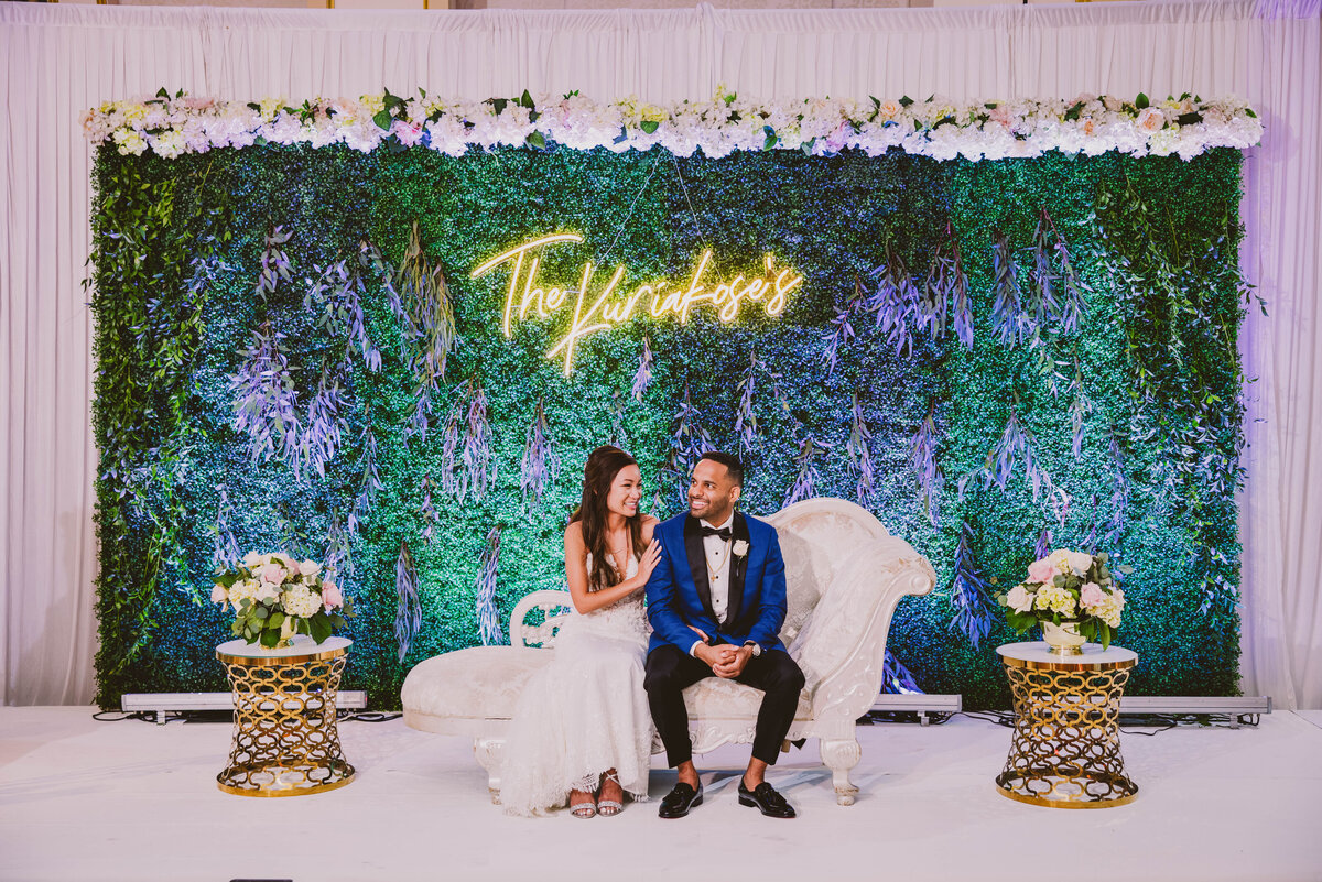 An indian christian bride and groom sitting on their wedding reception stage at the Grove NJ that has a green backdrop and an led sign with their name on it