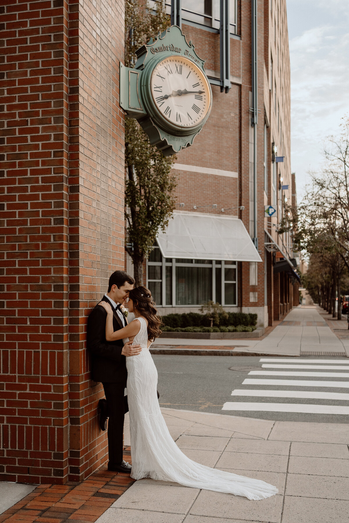 Bride and groom share a tender moment on a city sidewalk, leaning against a brick wall under a vintage clock. The bride, wearing an elegant white dress with a long train, embraces the groom, who is dressed in a black suit. The scene captures the romance and urban charm of their wedding day, set against the backdrop of city buildings and a crosswalk.