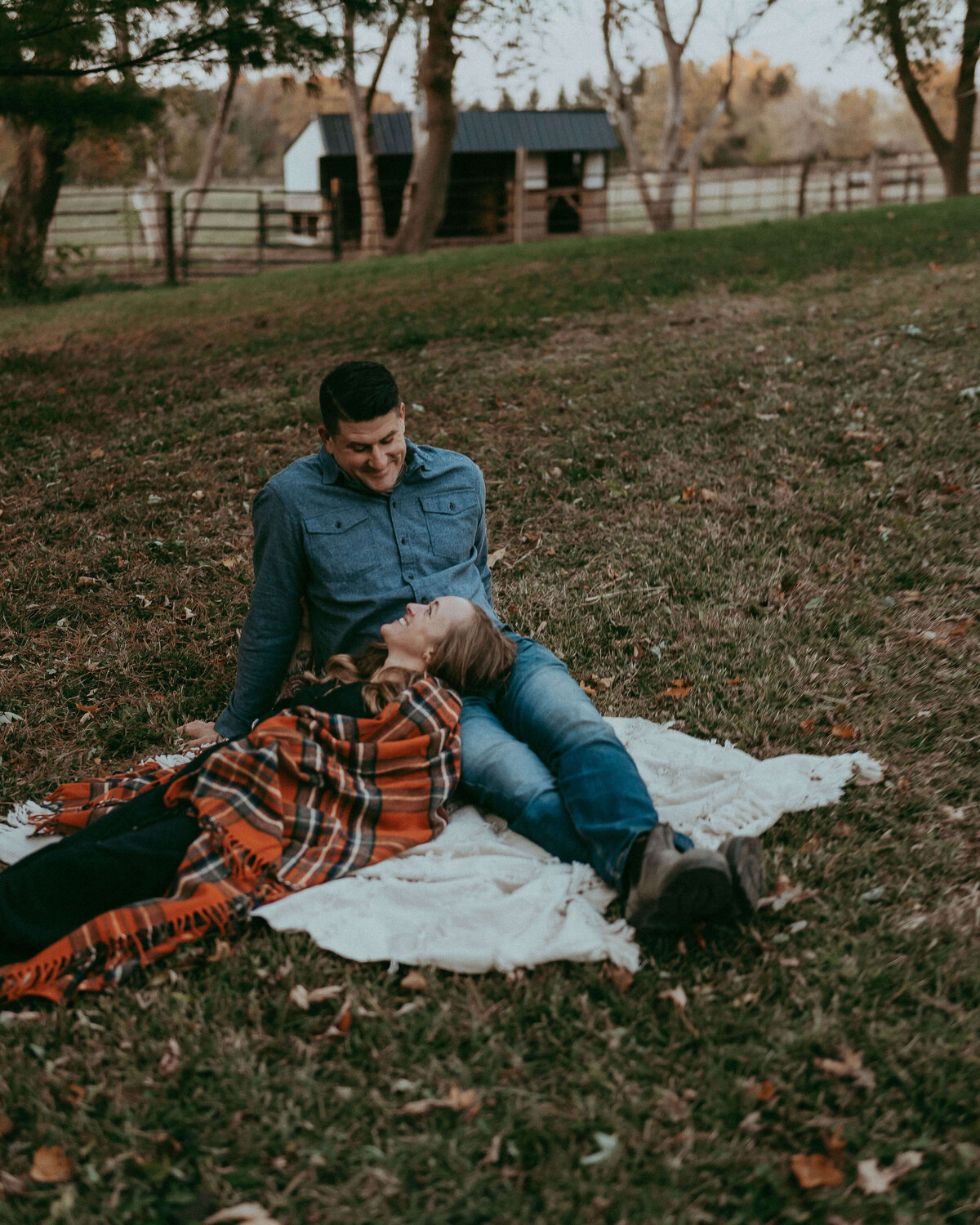 A wife cuddled up in a plaid blanket lays on her husband's lap while they sit outside of their family farm enjoying an autumn evening.