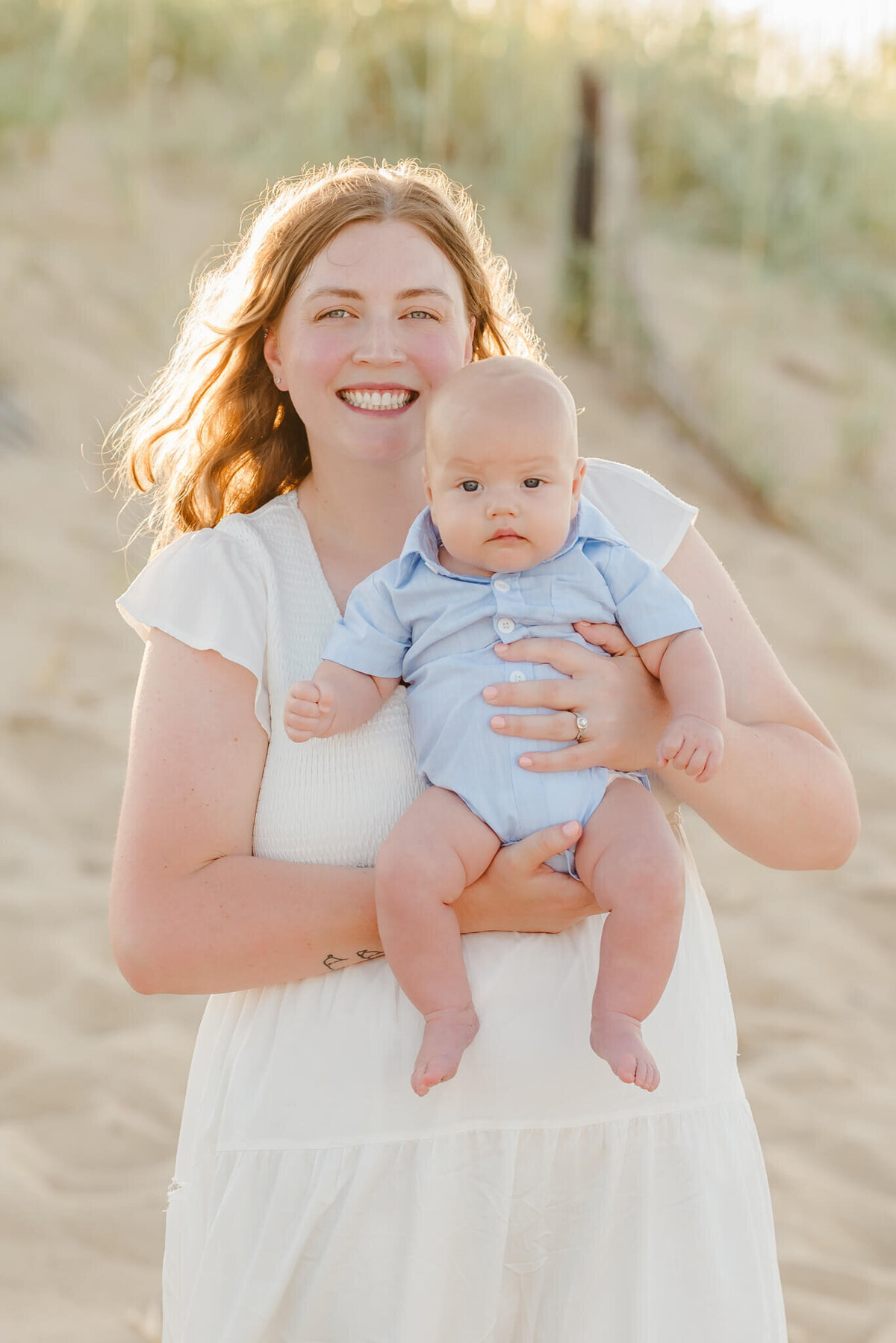 A mother in a white dress is holding her infant son, who is in a blue collared onesie. She is smiling big and the sun is lighting up her hair behind her.